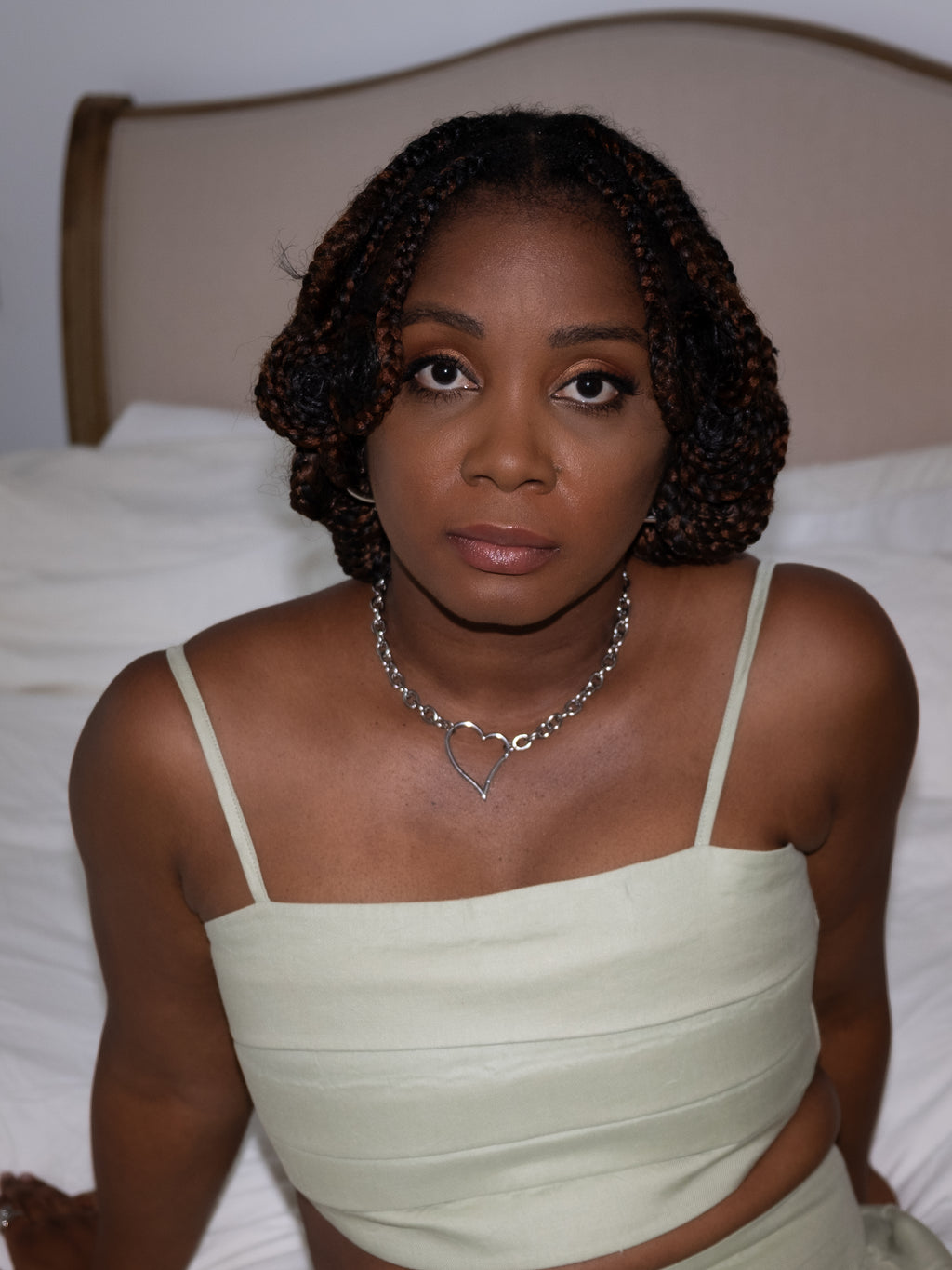 Woman wearing a light green dress with thin straps, sitting on a bed, wearing a necklace.